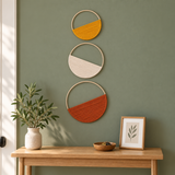 Three circular wall hangings with half-circle woven textile inserts in mustard yellow, off-white, and terracotta orange, displayed on a sage green wall above a light wood console table with a potted plant and framed botanical print.