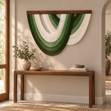 A modern wall-mounted art piece featuring layered, flowing green and off-white fabric-like loops suspended from a dark wood frame, displayed above a wooden console table in a warm, sunlit interior.