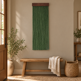 A green woven wall hanging with a dark wood top bar is displayed on a beige wall above a wooden bench in a styled entryway. A cream throw blanket is draped over the bench, with a woven basket and potted plants nearby.