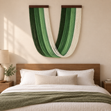 A large U-shaped wall hanging made of vertical woven stripes in shades of green and off-white, displayed above a neatly made bed in a warm, modern bedroom.