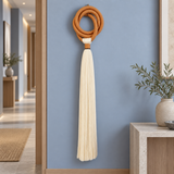 A decorative wall hanging with a knotted tan cord and a long cream tassel, mounted on a blue wall in a modern interior hallway with plants and a console table.