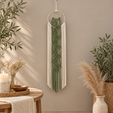 A green and white macrame wall hanging with a leaf pattern and long fringe, suspended from a wooden ring on a beige wall in a styled room with plants and a wooden table.