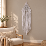 A white macrame wall hanging with a triangular top and long fringe, displayed on a beige wall in a softly lit room with a chair, throw blanket, and plant visible.