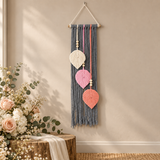 A bohemian-style woven wall hanging macrame wall decor with three hanging leaf-shaped ornaments in cream, pink, and coral, suspended from a wooden dowel on a beige wall next to a rustic wooden stand with flowers.