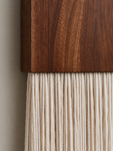 Close-up detail of a macrame wall hanging with a dark brown wood block and off-white cotton cords against a neutral wall.