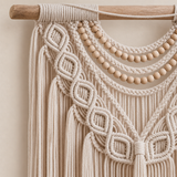 Cream macrame wall hanging with intricate knotted patterns, hanging from a wooden dowel with a row of round wooden beads, shown against a warm beige background.