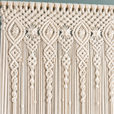 Cream macrame wall hanging with intricate woven patterns and long fringe, shown against a muted blue-gray background.