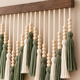 A close-up of a bohemian-style wall hanging featuring cream and sage green tassels, wooden beads, and a dark brown wooden top bar, mounted on a beige wall.