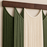 Macrame wall hanging with two green rope panels and one cream rope panel, suspended from a dark wood rod against a light beige wall.
