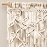 Cream macrame wall hanging with intricate knotted leaf patterns and long fringe, suspended from a light natural wood rod against a neutral beige background.