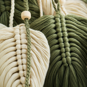 Close-up of woven macrame wall hangings in olive green and cream cotton cord, with a wooden bead accent on a hanging string.