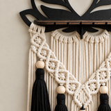 A close-up of a bohemian macrame wall hanging with cream-colored rope, black tassels, light wood beads, and a black wooden top bar mounted on a beige wall.
