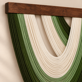 Close-up of a woven wall hanging with layered green and cream rope strands suspended from a dark wooden bar against a plain beige wall.