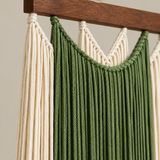 A close-up of a woven macrame wall hanging suspended from a dark wood rod. The hanging features thick braided cords in olive green and cream, arranged in vertical and curved panels against a plain light beige wall.