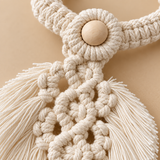 Close-up of a cream macrame wall hanging with a wooden bead at the top and long fringe tassels, set against a warm beige background.