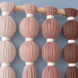 A close-up of a wall-hanging decorative textile made of vertical strands of yarn or cord gathered into rounded loops, hanging from a wooden rod with small gold hooks. The colors shown are soft blush pink, dusty rose, mauve, and dark brown, separated by white bands, against a muted blue-gray background.