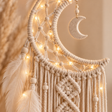 Cream macrame dreamcatcher wall hanging with woven rope, wooden beads, a crescent moon ornament, hanging feathers, and warm white string lights against a soft beige background.