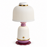 A decorative table lamp with a ribbed white shade and base, accented with gold trim, a pink neck, a purple base ring, and a scalloped dark purple footplate, shown on a white background.