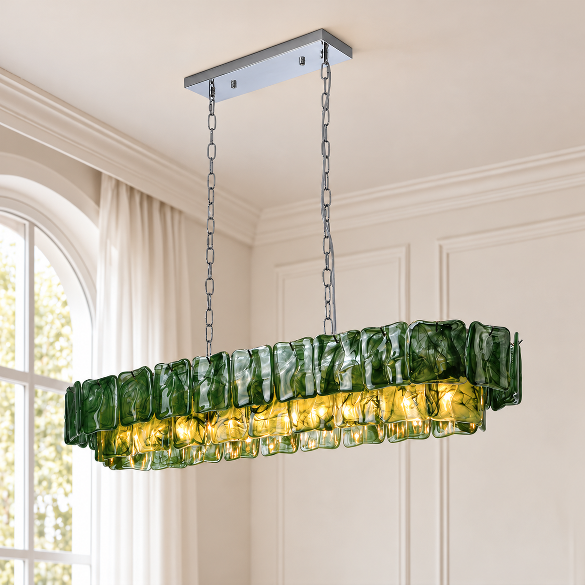 A linear chandelier with dark green rippled glass panels hangs from silver chains beside an arched window and cream paneled walls.