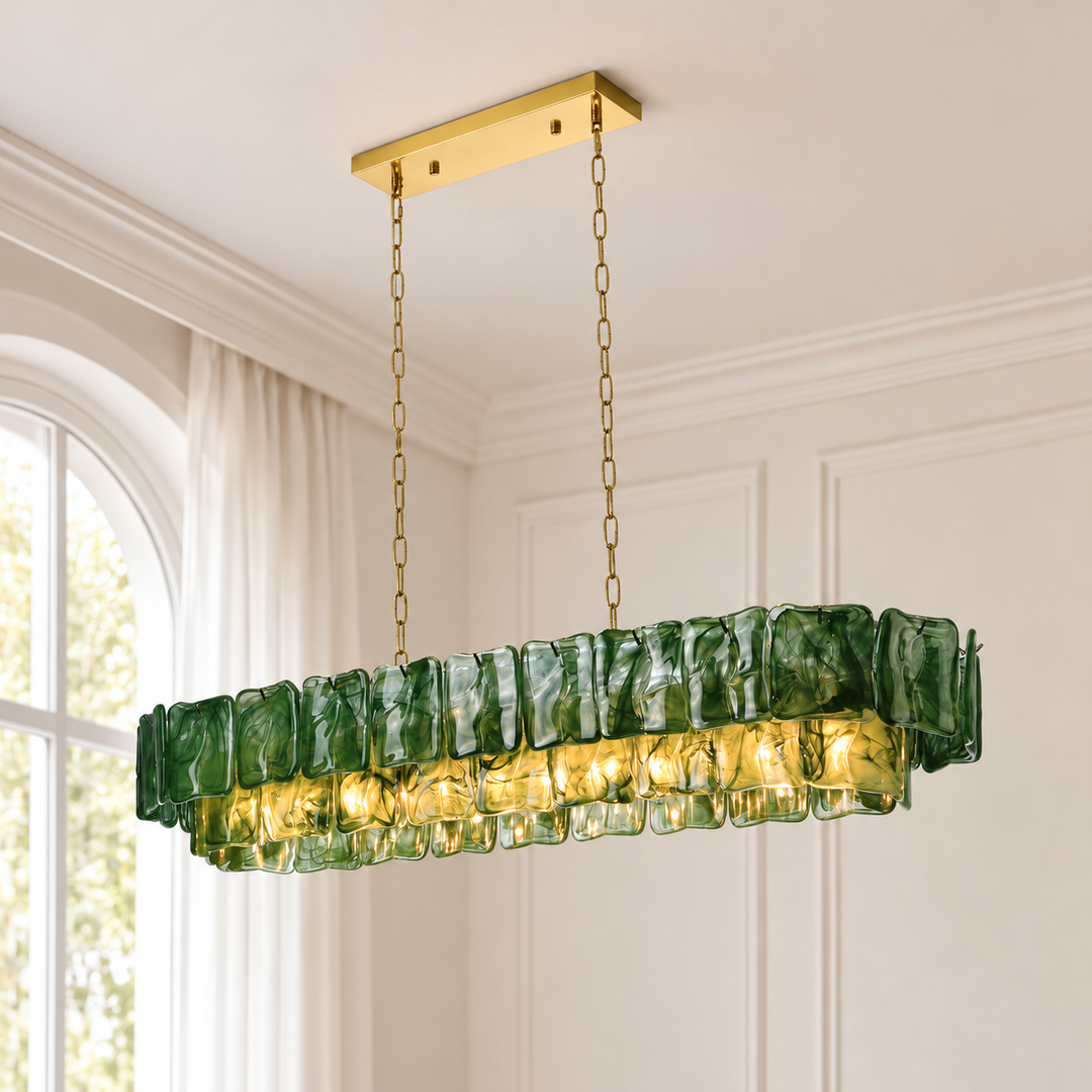 An illuminated dark green glass linear chandelier hangs from gold-toned chains against cream paneled walls beside an arched window.