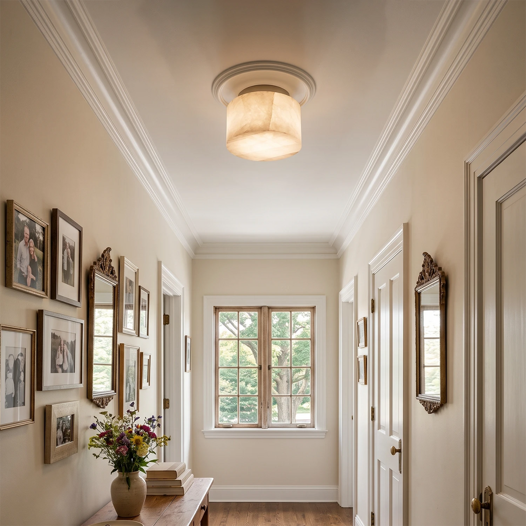 A warm, glowing flush-mount ceiling light with a drum shade in a classic hallway with white walls, framed pictures, and a window at the end.