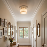 A warm, glowing flush-mount ceiling light with a drum shade in a classic hallway with white walls, framed pictures, and a window at the end.