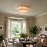 A modern flush-mount ceiling light with a textured, organic-shaped beige shade and a brushed silver metal rim, installed in a beautifully styled dining room with a round wooden table, white chairs, and a bay window.