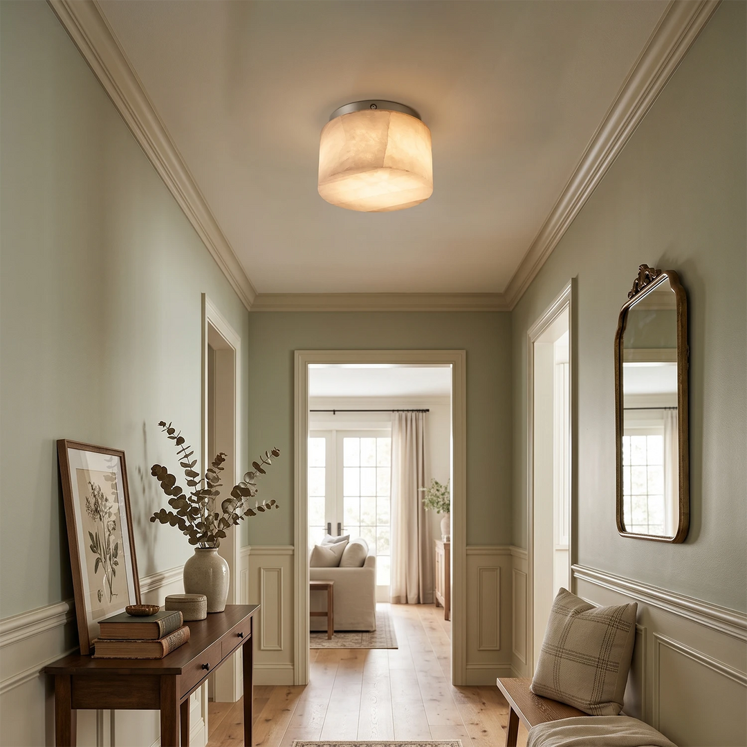 A warm, modern flush-mount ceiling light with a cylindrical fabric shade and brushed metal trim, glowing in a beautifully styled hallway with pale sage green walls, wood floors, and decorative accents.