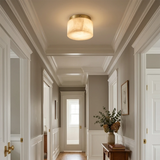 A warm, modern flush-mount ceiling light with a cylindrical drum shade and a brushed gold metal rim, mounted on a white coffered ceiling in a classic hallway with gray walls and white trim.