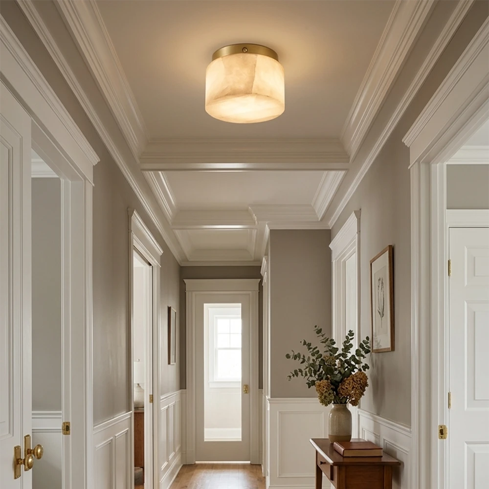 A warm, modern flush-mount ceiling light with a cylindrical drum shade and a brushed gold metal rim, mounted on a white coffered ceiling in a classic hallway with gray walls and white trim.