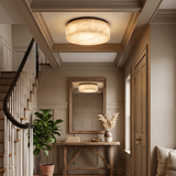 A modern flush-mount ceiling light with a textured, linen-like drum shade in warm cream and beige tones, glowing in a beautifully styled entryway with a staircase, console table, and mirror.
