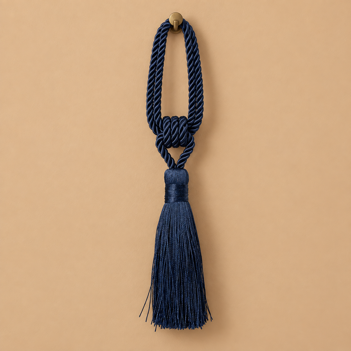 A navy blue decorative tassel with a knotted cord loop and a small round brass-tone finial, centered on a warm beige background.