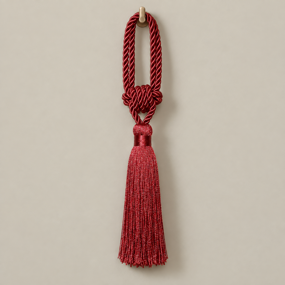 A red decorative tassel with a knotted cord loop and a glittery tassel, hanging from a small gold wall hook against a plain beige background.