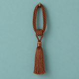 A brown braided rope tassel ornament with a knotted center and a long hanging tassel, shown against a solid teal background.