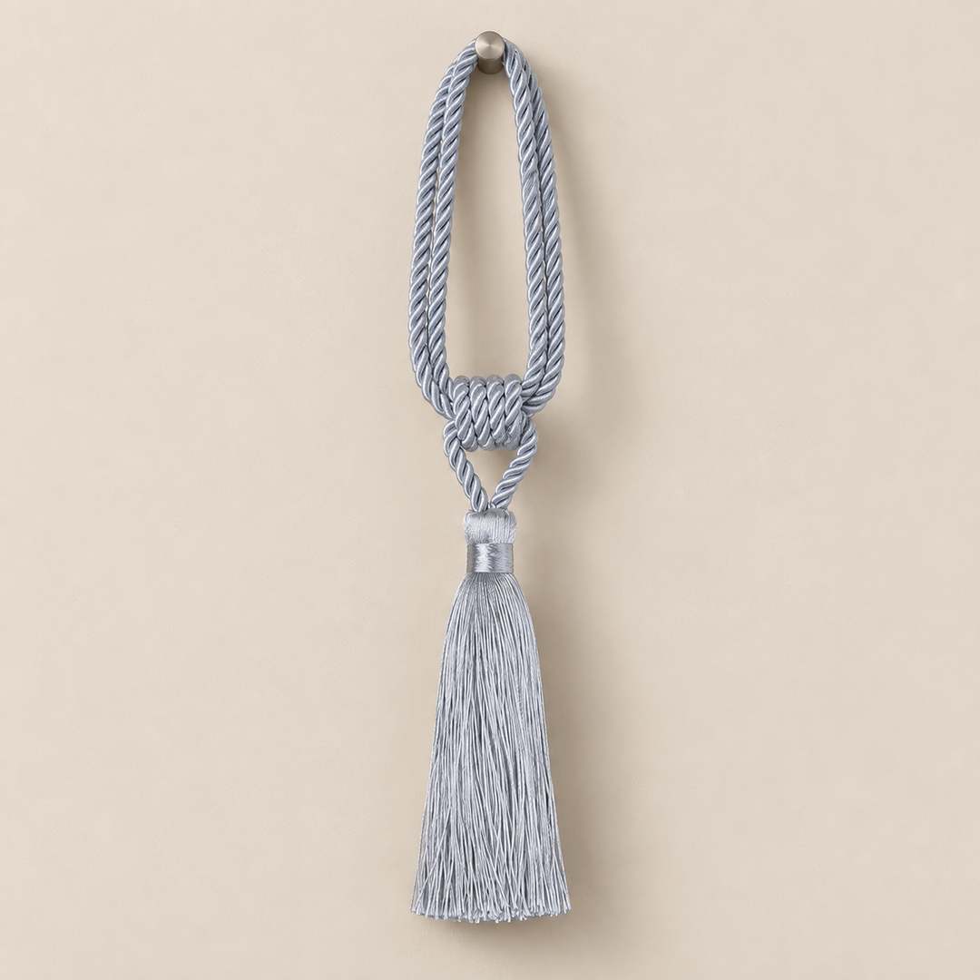A light blue-gray decorative tassel with a knotted cord loop and a silver-toned metal cap, shown against a solid beige background.