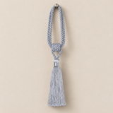 A light blue-gray decorative tassel with a knotted cord loop and a silver-toned metal cap, shown against a solid beige background.