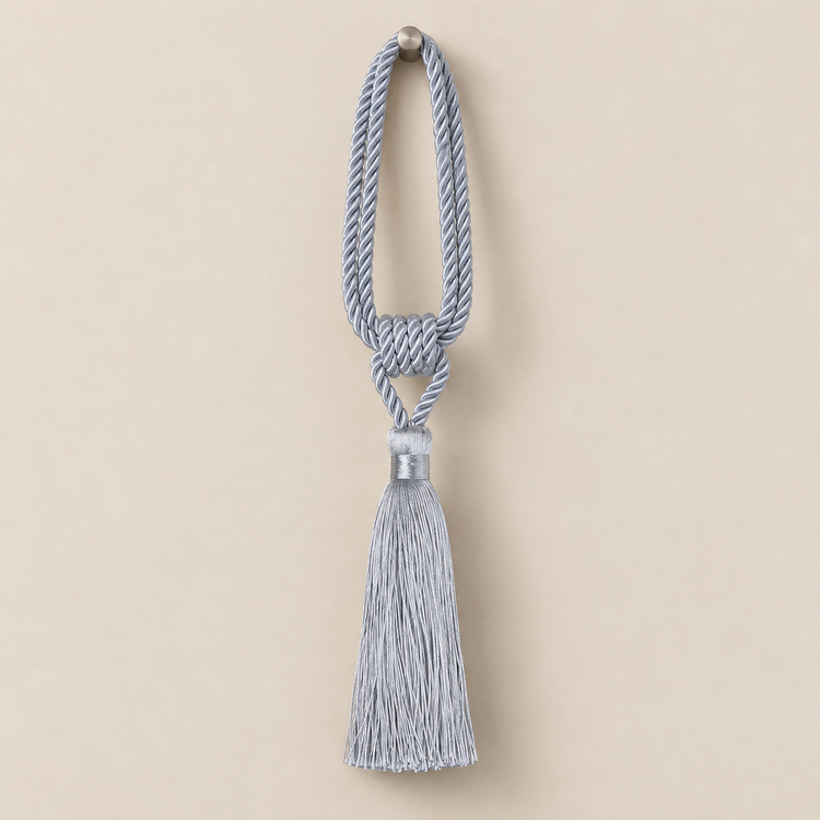 A light blue-gray decorative tassel with a knotted cord loop and a silver-toned metal cap, shown against a solid beige background.
