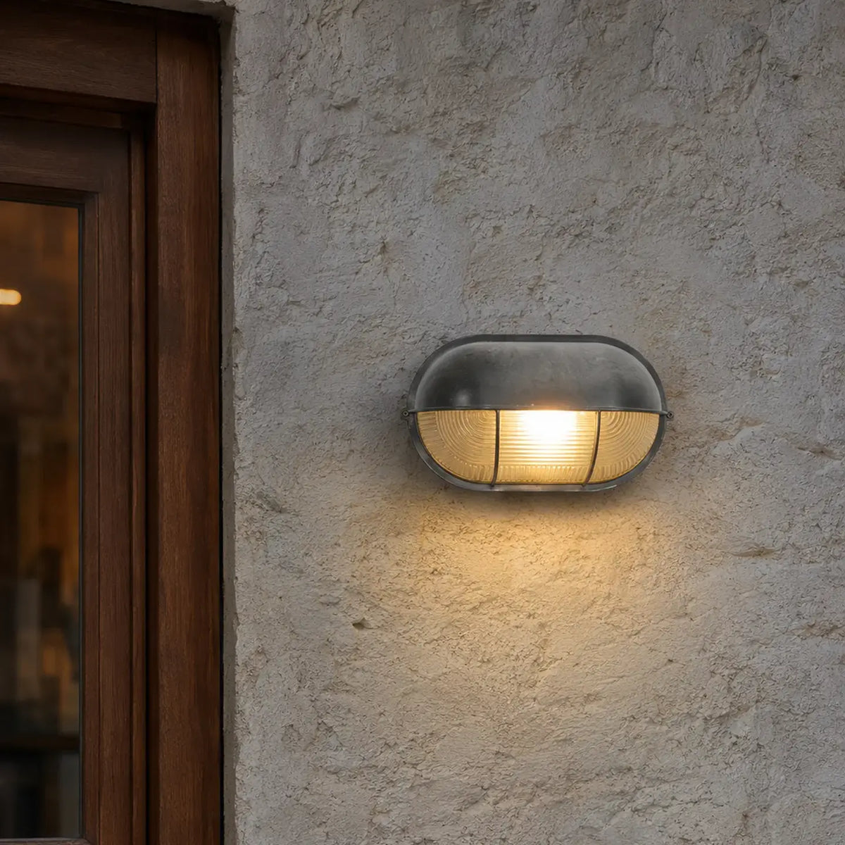 Oval silver metal wall sconce casting warm light down a textured gray wall beside a dark wooden window frame.