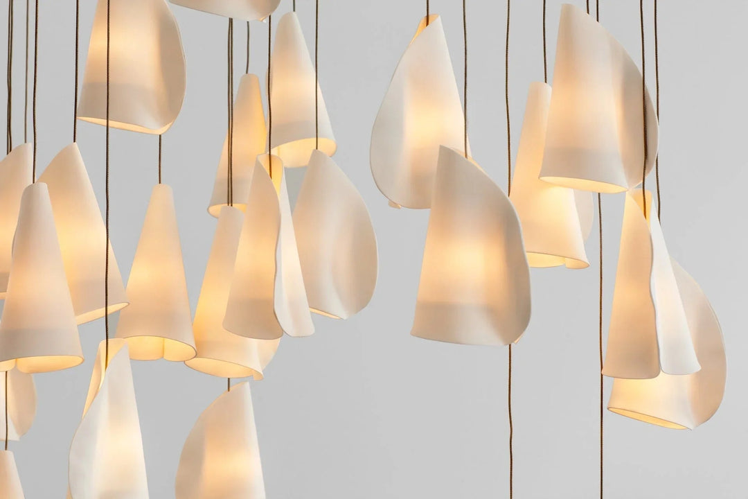 Chandelier with glowing white porcelain folded shades suspended at varied heights on dark cords against a light gray background.