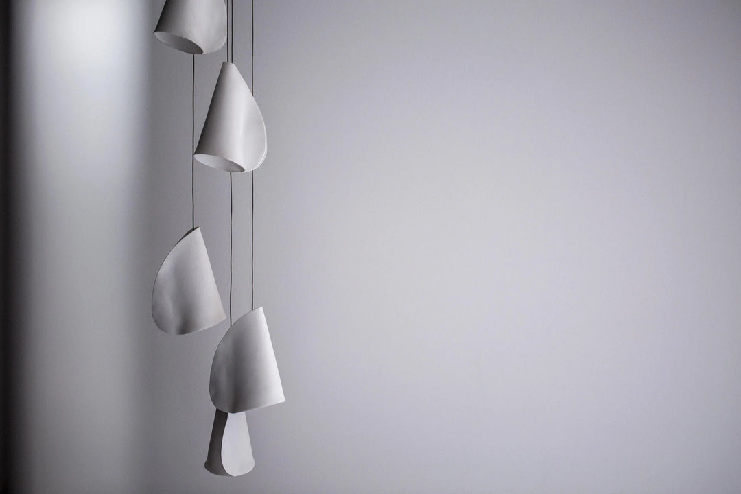 Unlit white chandelier shades hang vertically at staggered heights on thin dark cords against a gray wall.