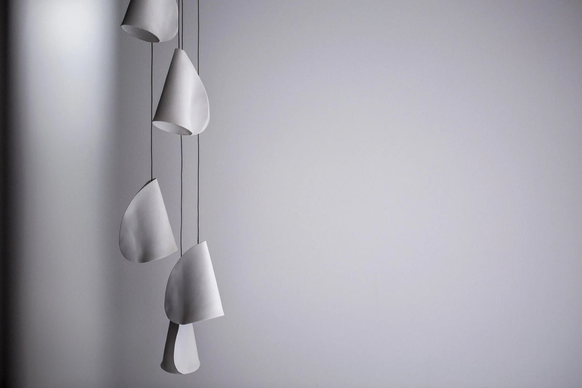 Unlit white chandelier shades hang vertically at staggered heights on thin dark cords against a gray wall.