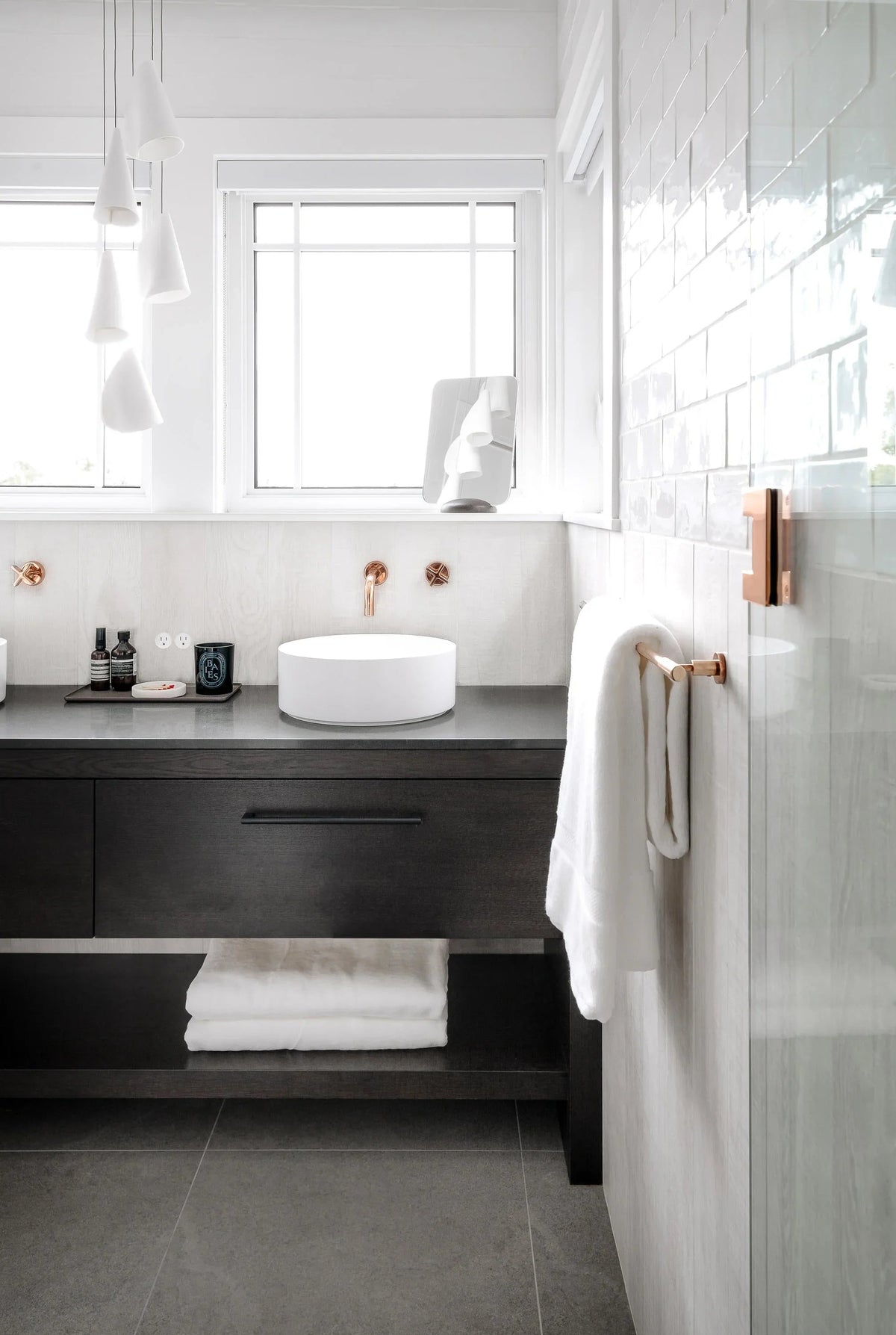 White folded-shade chandelier hangs beside bathroom windows above a dark vanity with a round white basin.