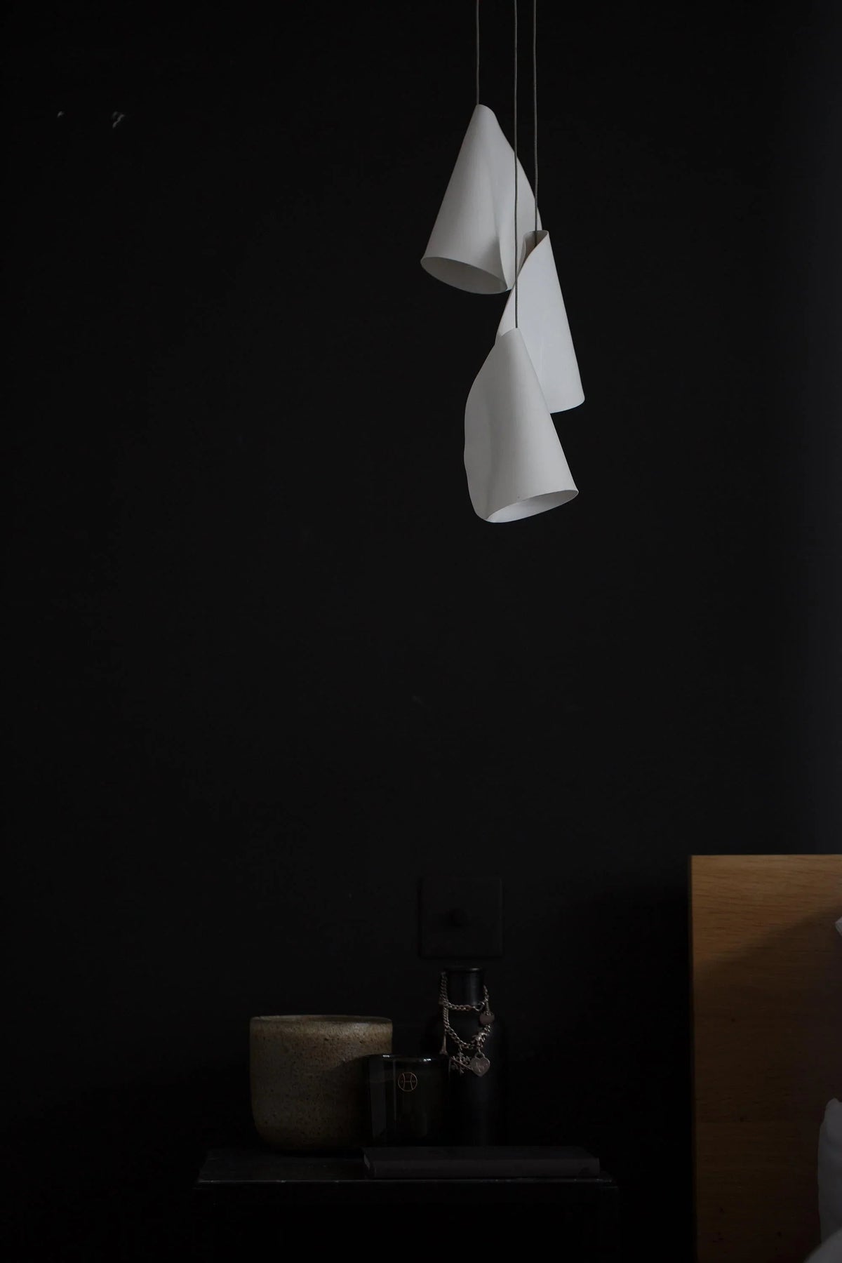 Three white folded chandelier shades hang at staggered heights against a black wall above a small side table.