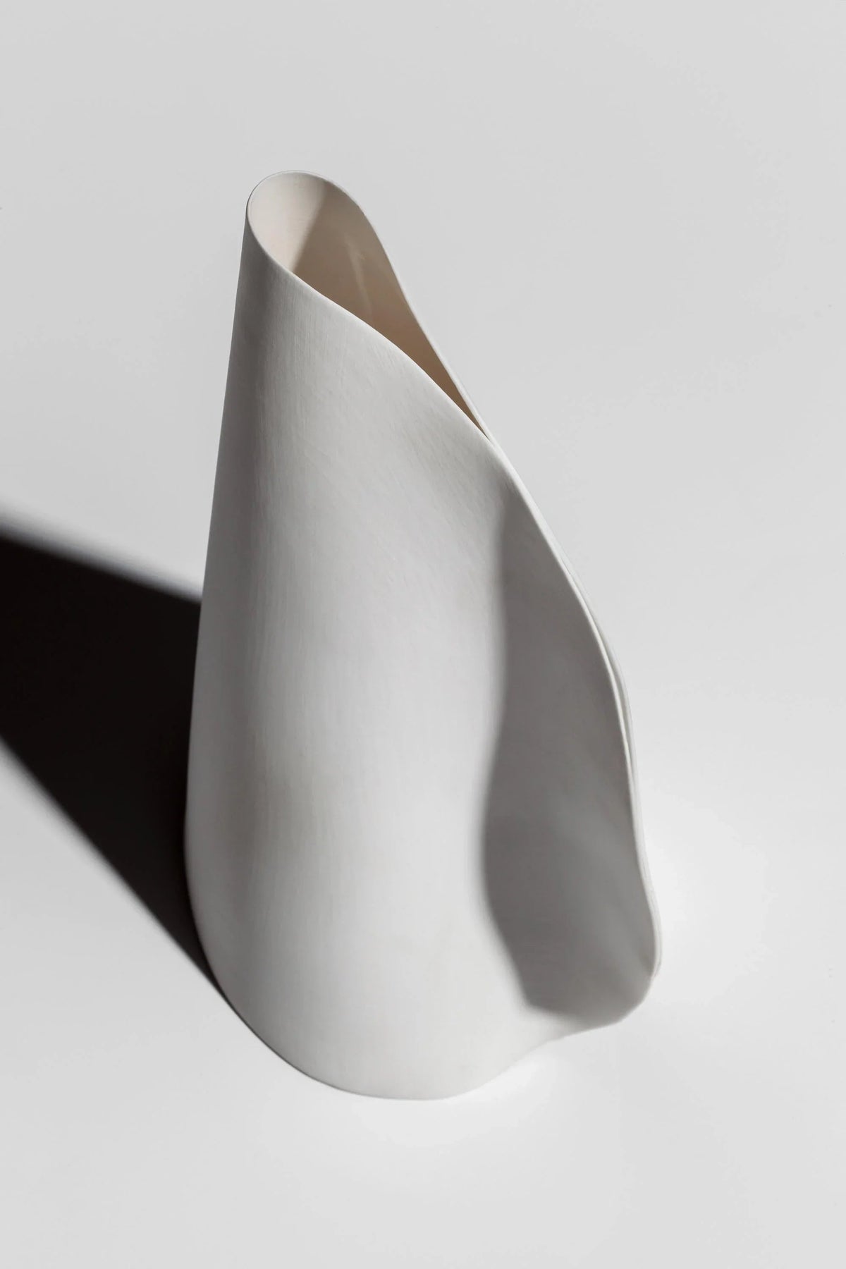 A white porcelain chandelier shade stands upright on a pale surface, showing its curved folded form and casting a dark shadow.