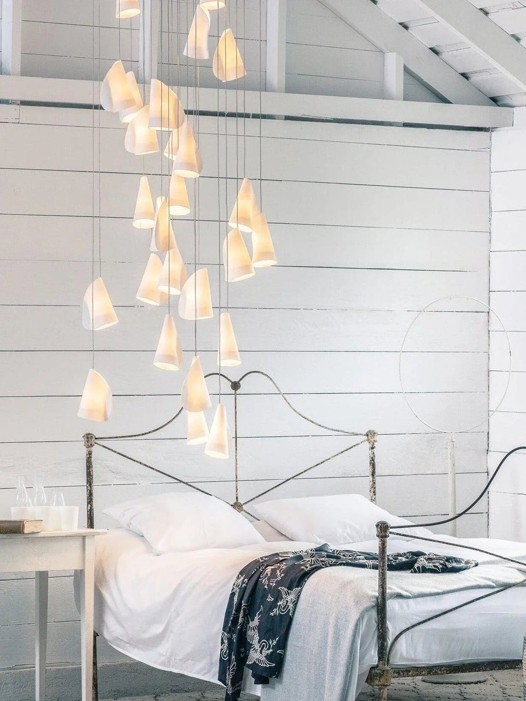 White porcelain chandelier cascades above a metal bed frame against white plank walls beneath exposed ceiling beams.