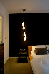 Five glowing white porcelain shades form a cascading chandelier beside a wooden bed against a black bedroom wall.