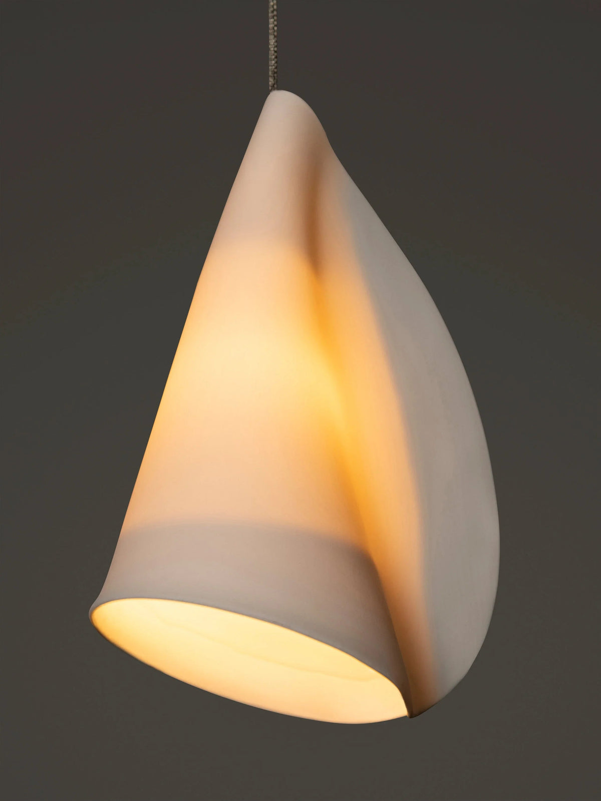 Close-up of a chandelier’s folded white porcelain shade glowing warmly against a dark grey background.