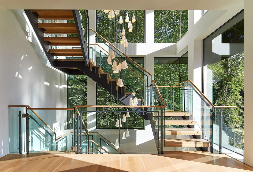White porcelain chandelier cascading through an open stairwell with wooden steps, glass railings, and large windows overlooking greenery.