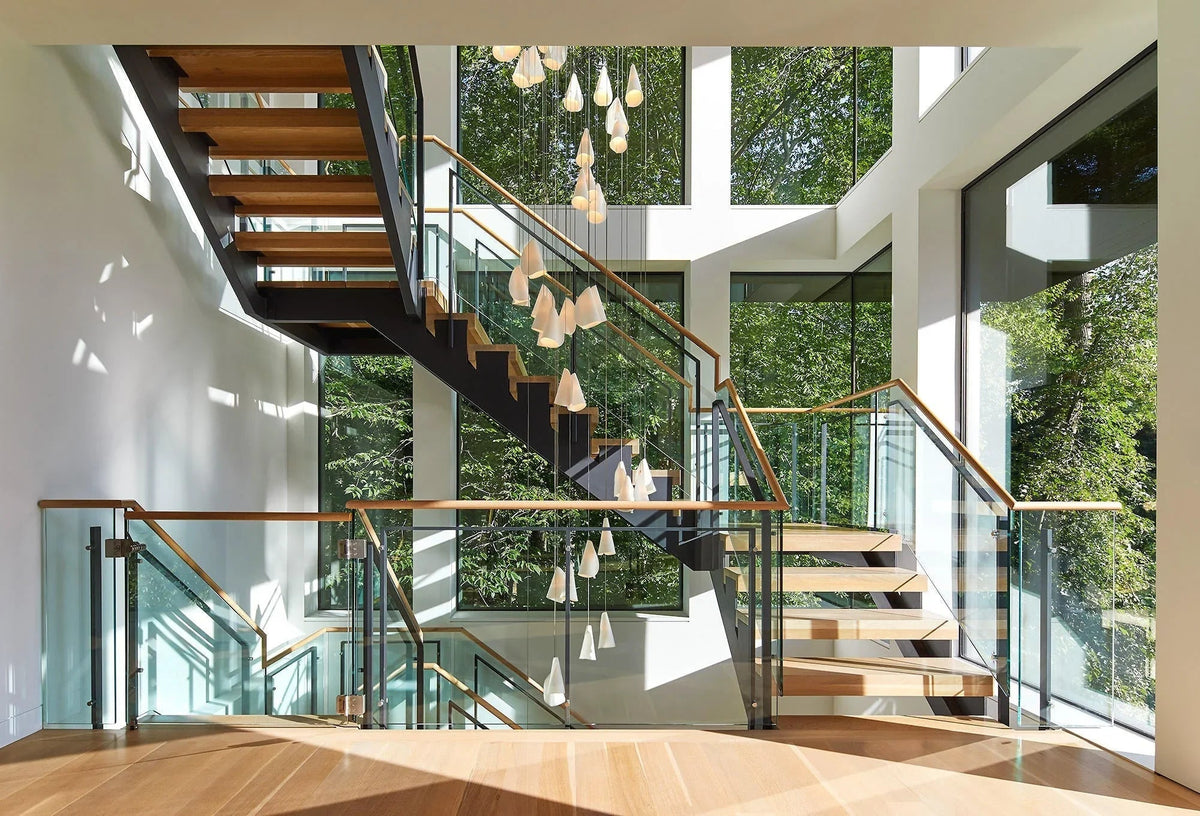 White porcelain chandelier cascading through an open stairwell with wooden steps, glass railings, and large windows overlooking greenery.