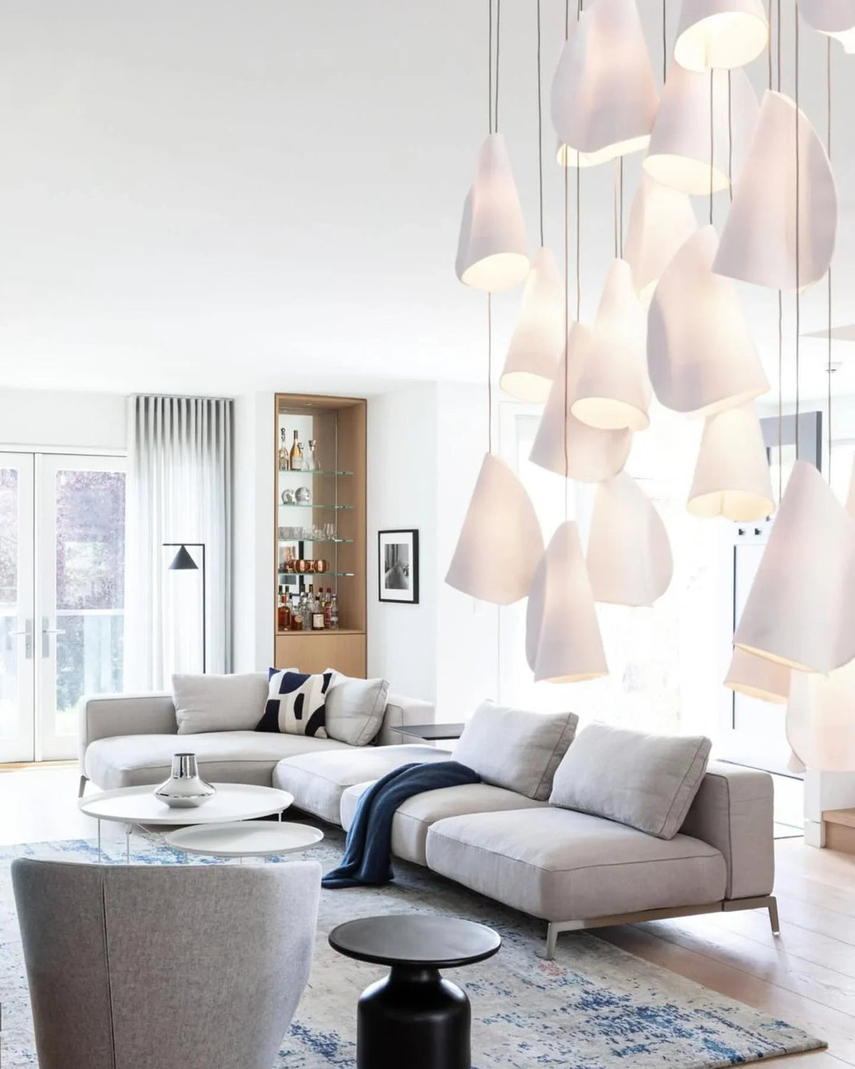 Glowing white porcelain chandelier fills the foreground of a bright living room with a pale sectional sofa and round tables.